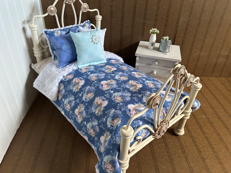 May include: A miniature white metal bed with a blue comforter featuring a princess and snowflake pattern. The bed is made up with blue and light blue pillows. A white nightstand with two drawers is next to the bed.