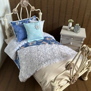 May include: Miniature white metal bed with a cream-colored mattress and bedding. The bed is adorned with blue patterned pillows and a light blue pillow with a snowflake design. A small white nightstand with a vase of flowers is next to the bed.