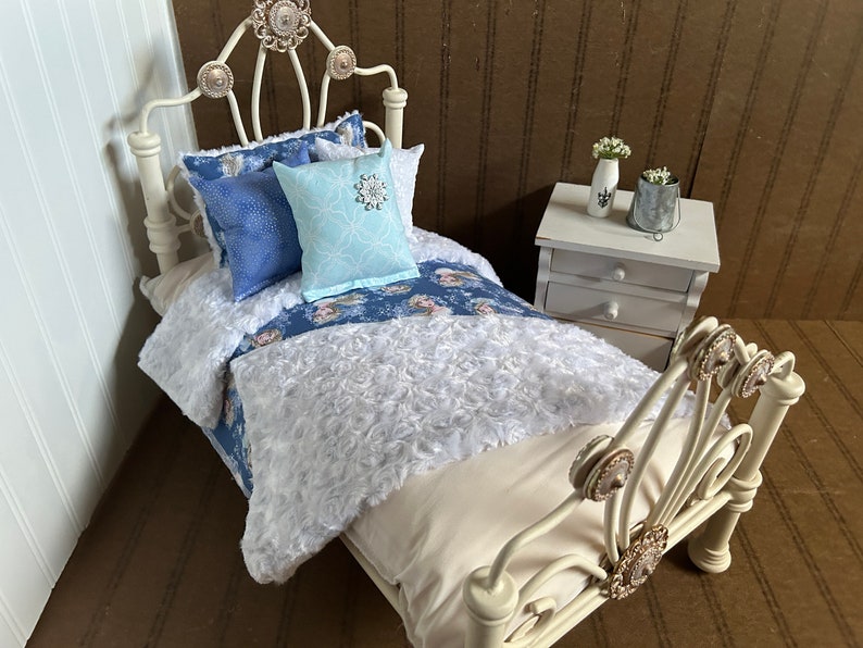 May include: Miniature white metal bed with a cream-colored mattress and bedding. The bed is adorned with blue patterned pillows and a light blue pillow with a snowflake design. A small white nightstand with a vase of flowers is next to the bed.