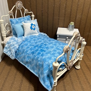 May include: A miniature white metal bed with a blue floral patterned comforter, pillows, and a white nightstand. The bed frame has decorative accents. The bedding includes a blue floral pattern and a white pillow with a blue flower.