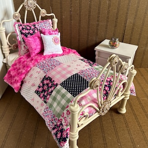 May include: A miniature doll bed with a cream-colored metal frame and a patchwork quilt. The quilt features pink, white, and navy blue floral and polka dot patterns. The bed is adorned with pink pillows and a fluffy pink blanket.