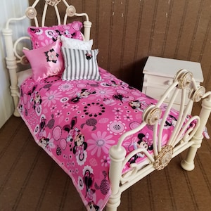 May include: A white metal doll bed with a pink and black floral bedding set featuring Minnie Mouse. The bed has a decorative headboard and footboard with gold accents.