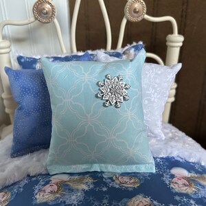 May include: A doll-sized bed with blue and white bedding. The bed has a white metal frame with decorative knobs. Pillows include a light blue pillow with a snowflake and patterned pillows.