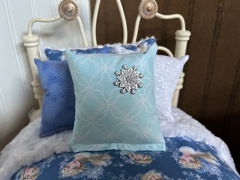 May include: A doll-sized bed with blue and white bedding. The bed has a white metal frame with decorative knobs. Pillows include a light blue pillow with a snowflake and patterned pillows.