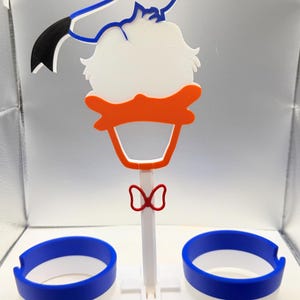 May include: A 3D printed plastic stand featuring a stylised white duck head with an orange beak and a blue sailor hat. The stand includes a central white post with a red bow tie and two circular blue and white cup holders at the base.