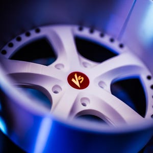 May include: A white five-spoke wheel rim featuring a circular red center cap with the gold letters Vs printed in the middle. The rim has a metallic silver outer barrel with a series of small circular bolt holes along the inner edge of the spokes.