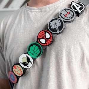 May include: A gray fabric strap featuring a series of circular, raised icons representing various superhero symbols, including a star, a helmet, a claw mark, a green fist, a spider mask, a hammer, a robot face, and an Avengers logo.