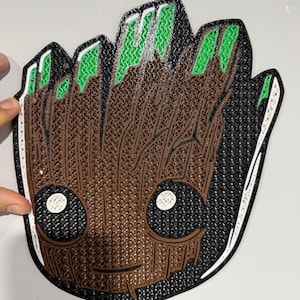 May include: A 3D printed wall art piece featuring a stylized character with a brown wood-grain texture, large black eyes with white pupils, and green accents on the top. The design is outlined in black with a white border on a flat surface.