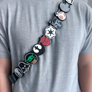 Op de afbeelding: Een zwarte schouderband versierd met een reeks ronde en gevormde siliconen bedels met diverse Star Wars-personages en symbolen, waaronder Darth Vader, Grogu, het keizerlijke embleem en een Stormtrooper-helm, gedragen over een grijs T-shirt.