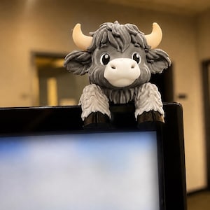 Könnte beinhalten: Eine kleine, graue, dreidimensionale Highland-Rind-Figur mit weißer Schnauze, cremefarbenen Hörnern und schwarzen Hufen. Die Figur hat ein strukturiertes, zotteliges Fell und ist so gestaltet, dass sie auf dem oberen Rand eines Monitors sitzt.