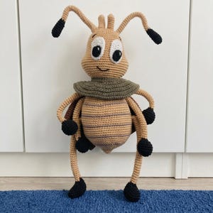 May include: A tan crochet insect toy with a round head, large white eyes with black pupils, and two antennae with black tips. It features a dark olive green collar, a striped tan and brown body, and six long limbs ending in black crochet feet.