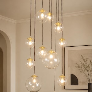 Può includere: Un lampadario a più luci caratterizzato da un perno centrale dorato con bracci radianti. Dodici globi sferici in vetro trasparente di varie dimensioni pendono ad altezze diverse da cavi scuri intrecciati, ciascuno contenente una lampadina.