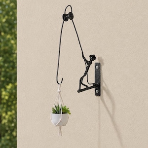 Pode incluir: Um suporte de parede para plantas em metal preto com uma figura estilizada puxando uma corda sobre uma roldana, com um gancho na extremidade que segura um pequeno vaso de cerâmica branca com uma planta suculenta verde em macramé.