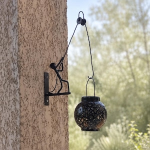 Könnte beinhalten: Eine schwarze Wandhalterung aus Metall mit einer stilisierten Figur, die einen dünnen Draht hält, an dem eine kugelförmige Laterne hängt. Die Laterne hat eine dunkle Oberfläche mit filigranen Blumenmustern, die Licht durchlassen.