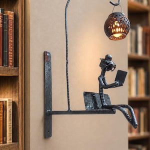Peut inclure: Une applique murale en métal noir représentant une silhouette portant un chapeau, assise sur un banc et lisant un livre. Un bras arqué s'étend du mur, soutenant un luminaire sphérique perforé qui projette une lueur chaude vers le bas.