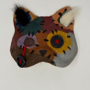 May include: A cat-shaped patch made of multi-colored felt pieces in brown, yellow, and grey, joined by black whipstitching. It features two black buttons as eyes, one attached with red thread, and tufts of black and white fur on the ears.