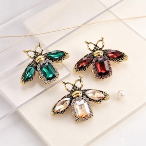 Peut inclure: Trois broches en forme d'abeille en métal doré vieilli, chacune ornée d'une grande pierre rectangulaire centrale, d'ailes en forme de marquise et d'un contour de minuscules strass transparents. L'ensemble comprend une broche verte, une rouge et une champagne, accompagnées d'une petite perle blanche.