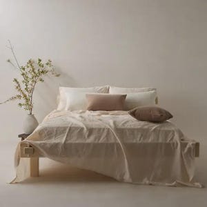 May include: A wooden bed frame with a light beige linen sheet, two white rectangular pillows, one brown rectangular pillow, and one brown square pillow. A white vase with thin branches containing small green leaves sits beside the bed.
