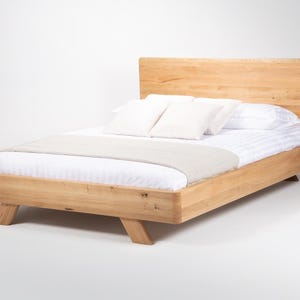 May include: A light-colored wooden bed frame with a rectangular headboard and angled legs. The bed is dressed with a white striped mattress cover, two white square pillows, and a light beige runner draped across the foot of the bed.