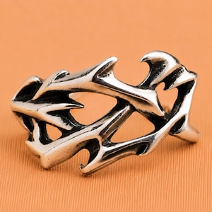 Peut inclure: Une bague en métal argenté présentant un design ajouré avec des protubérances pointues en forme d'épines et des bords dentelés. La surface du métal a une finition polie avec des zones en creux assombries pour souligner la forme angulaire.
