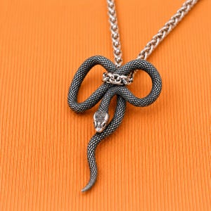 May include: A silver-toned metal necklace featuring a pendant shaped like a coiled snake with a textured, scale-like pattern. The snake's body loops to form a knot-like design, and it is attached to a matching silver-toned chain link necklace.