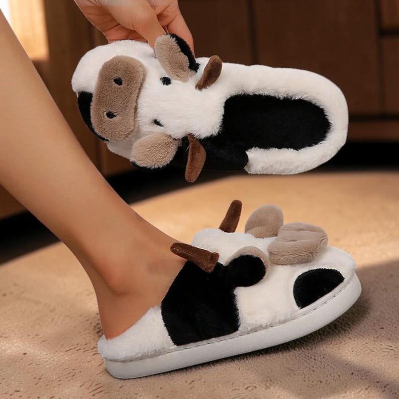 May include: A pair of plush, fuzzy slippers designed to resemble cows. The slippers feature a white base with black patches, brown snout details, and small brown ears. The interior is lined with black fabric, and the soles are white and flat.