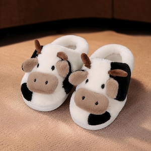 May include: A pair of plush, white, black, and tan cow-themed slippers. Each slipper features a soft, fuzzy texture, a raised tan snout, small brown ears, and black spots. The design includes a rounded toe and a white sole, mimicking the appearance of a cow's face with embroidered eyes and nostrils.