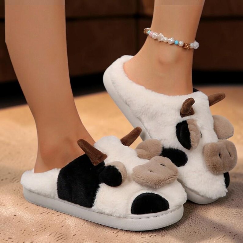 May include: A pair of plush, white and black cow-themed slippers featuring soft faux fur, brown fabric horns, and a light brown snout. The slippers have a slip-on design with a rounded toe and a flat, light gray sole, showcasing a whimsical animal pattern.
