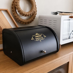 May include: A matte black metal bread box with a roll-top lid and a silver knob. The front features gold decorative filigree and the text "The Vintage BREAD" printed in a stylized font. The container has a rectangular base with rounded edges.