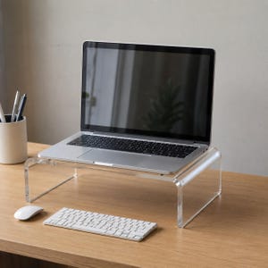 Może przedstawiać: Przezroczysta podstawka pod laptopa z akrylu o kształcie litery U, podtrzymująca srebrny laptop z czarnym ekranem. Podstawka znajduje się na drewnianej powierzchni, a przed nią umieszczono białą klawiaturę bezprzewodową oraz białą mysz komputerową.