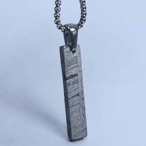May include: A rectangular pendant with a dark grey, textured, metallic surface featuring a distinct cross-hatched pattern, suspended from a silver-toned metal chain. The pendant has a smooth, polished bail connecting it to the link chain.