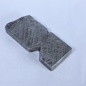 May include: A rectangular iron meteorite slice featuring a distinct Widmanstätten pattern of intersecting metallic lines. The surface is dark gray with a metallic sheen, showing a complex geometric crystalline structure across the entire face.