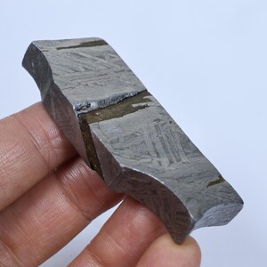 May include: A metallic meteorite fragment with a distinct Widmanstätten pattern of intersecting crystalline lines across its grey surface, featuring a dark, recessed diagonal fissure that cuts across the centre of the rectangular specimen.