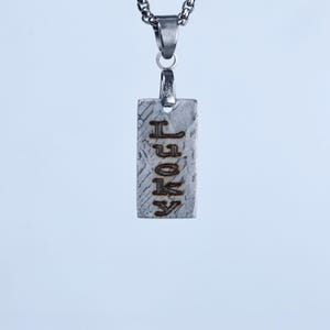 Puede incluir: Un colgante rectangular de metal plateado que cuelga de una cadena a juego. El colgante presenta una superficie texturizada y desgastada con la palabra LUCKY grabada verticalmente en letras mayúsculas oscuras y negritas. El metal brilla.