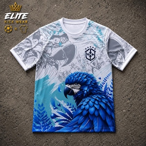 Puede incluir: Una camiseta de manga corta con cuello redondo que presenta un gráfico de un guacamayo azul vibrante, un fondo abstracto en blanco y azul claro, y un escudo CBF azul oscuro en el pecho. En la esquina superior izquierda aparece el texto ELITE KICK WEAR.