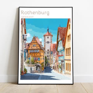 May include: A framed wall art print featuring a colourful illustration of a street in Rothenburg, Germany. The image displays half-timbered buildings in yellow and blue, a tall stone clock tower, and a cobblestone path under a bright blue sky.