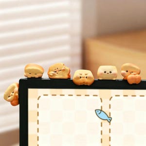 Puede incluir: Un conjunto de seis pequeñas figuras decorativas de color marrón claro con forma de pan y caras de dibujos animados, posadas en el borde superior de un marco de monitor negro. Cada figura muestra una expresión única, incluyendo sonrisas.