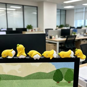 May include: A set of six small banana-shaped figurines with yellow peels and white bodies, each featuring a simple face with two eyes and a small mouth. The figures are posed in various positions along the top edge of a black computer monitor.