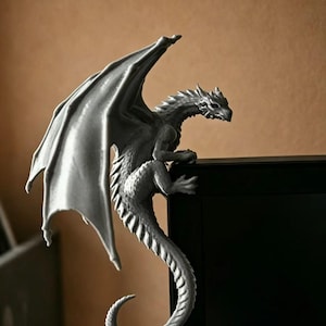 May include: A grey, sculpted dragon figurine with detailed scales and large, outstretched wings. The creature is perched on the top corner of a black rectangular object, with its long, curved tail extending downwards along the side of the frame.