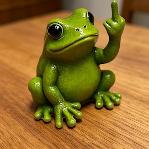 Könnte beinhalten: Eine kleine, hellgrüne Froschfigur mit großen, glänzenden schwarzen Augen. Der Frosch sitzt in einer aufrechten Position, wobei der rechte Arm erhoben ist und der Mittelfinger nach oben zeigt. Die Oberfläche der Figur ist glatt und poliert.