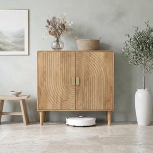 May include: A light brown wooden cabinet with two doors featuring a carved arch pattern and vertical lines. The cabinet has four tapered legs, two gold-toned oval handles, and a white robotic vacuum cleaner positioned on the floor underneath it.