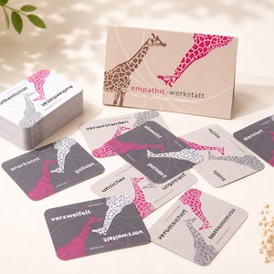 Puede incluir: Un mazo de cartas y una caja etiquetada como empathie-werkstatt con ilustraciones de jirafas en rosa y gris. Las cartas muestran varias palabras en alemán como authentizität, verstanden y verzweifelt, con patrones de jirafas.
