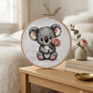 Peut inclure: Un tambour à broder circulaire en bois contenant un motif au point de croix représentant un koala gris assis. Le koala a de grands yeux noirs, l'intérieur des oreilles rose et des coussinets roses, et tient une petite fleur rose dans ses pattes.