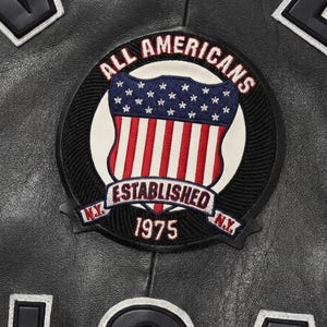 May include: A black leather jacket featuring a circular embroidered patch with the text ALL AMERICANS, ESTABLISHED, and 1975. The patch displays a shield with a blue field of white stars and red and white vertical stripes, flanked by N.Y. text.