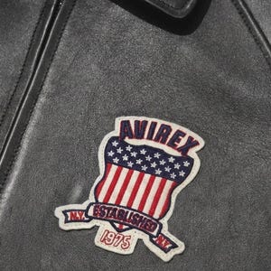 May include: A black leather jacket featuring a stitched patch with the text AVIREX, ESTABLISHED 1975, and N.Y. The patch displays a shield design with a blue field of white stars above red and white vertical stripes, all bordered in white.