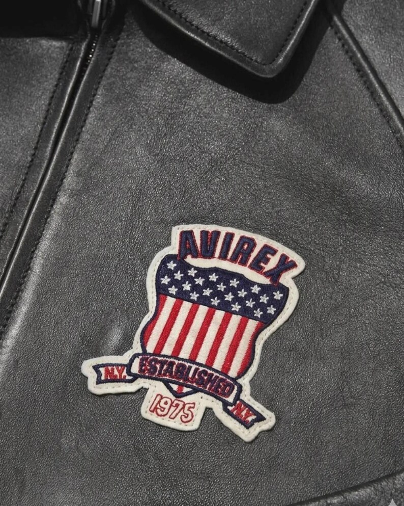 May include: A black leather jacket featuring a stitched patch with the text AVIREX, ESTABLISHED 1975, and N.Y. The patch displays a shield design with a blue field of white stars above red and white vertical stripes, all bordered in white.