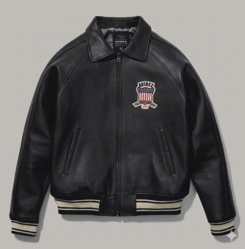 May include: A black leather jacket featuring a full-length front zipper, a classic collar, and long sleeves. The chest displays an embroidered patch with the text AVIREX above an American flag design. The cuffs and hem have ribbed striped trim.