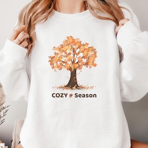Könnte beinhalten: Ein weißes langärmeliges Sweatshirt mit Rundhalsausschnitt und einer Grafik eines Baumes mit orangefarbenen und gelben Herbstblättern. Unter dem Baum steht der Text COZY SEASON in dunkelbrauner Schrift mit einem kleinen Eichelsymbol.
