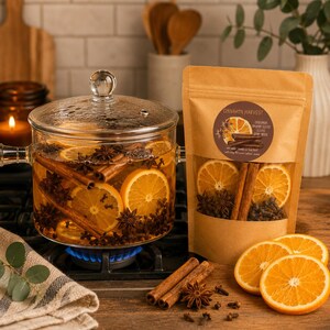 May include: A clear glass pot with a lid containing water, orange slices, cinnamon sticks, and star anise on a stovetop. Next to it is a brown paper pouch labeled Cinnamon Harvest with a window showing dried orange slices, cinnamon, and cloves.