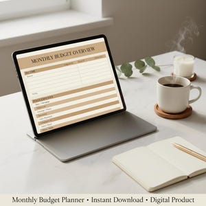 以下が含まれることがあります： Monthly Budget Overviewの表計算が表示されたタブレット、木製コースターに乗った白い陶器のコーヒーマグ、金色のペンが置かれた開いたノートが白い表面に配置されています。